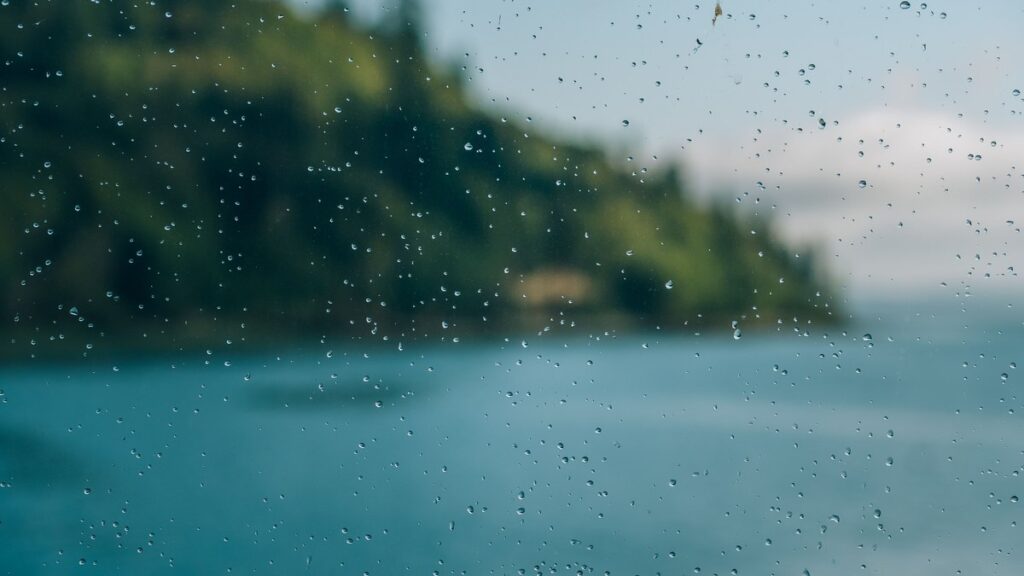 Regentropfen an einer Fensterscheibe, dahinter ein See mit bewaldetem Ufer