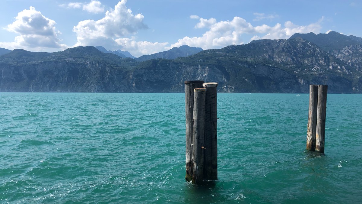 Klares türkisfarbenes Wasser des Gardasees mit alten Anlegepfählen, dahinter die Steilhänge des Ufers