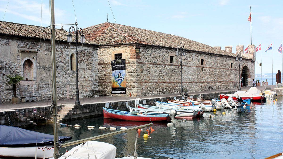 Langgestrecktes Steingebäude der Dogana Veneta am Hafen von Lazise, davor liegen bunte Ruderboote