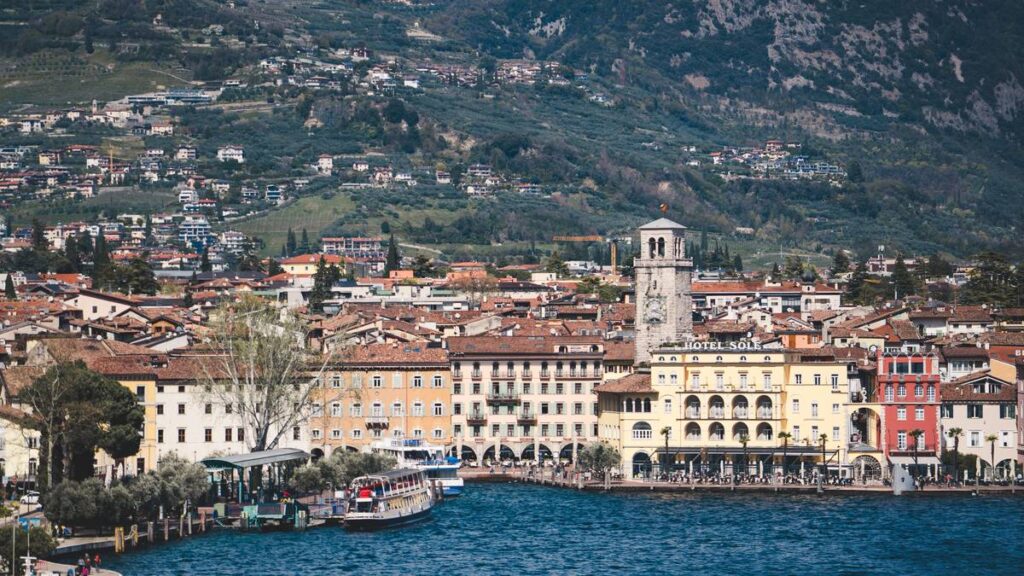 Die Uferpromenade von Riva del Garda mit der Torre Apponale, den Arkaden am Wasser und den Bergen des noerdlichen Gardasees