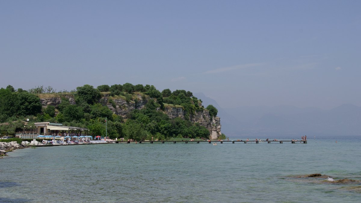 Flache Badestelle mit langem Holzsteg am Ufer der Halbinsel von Sirmione, dahinter eine bewachsene Felskante