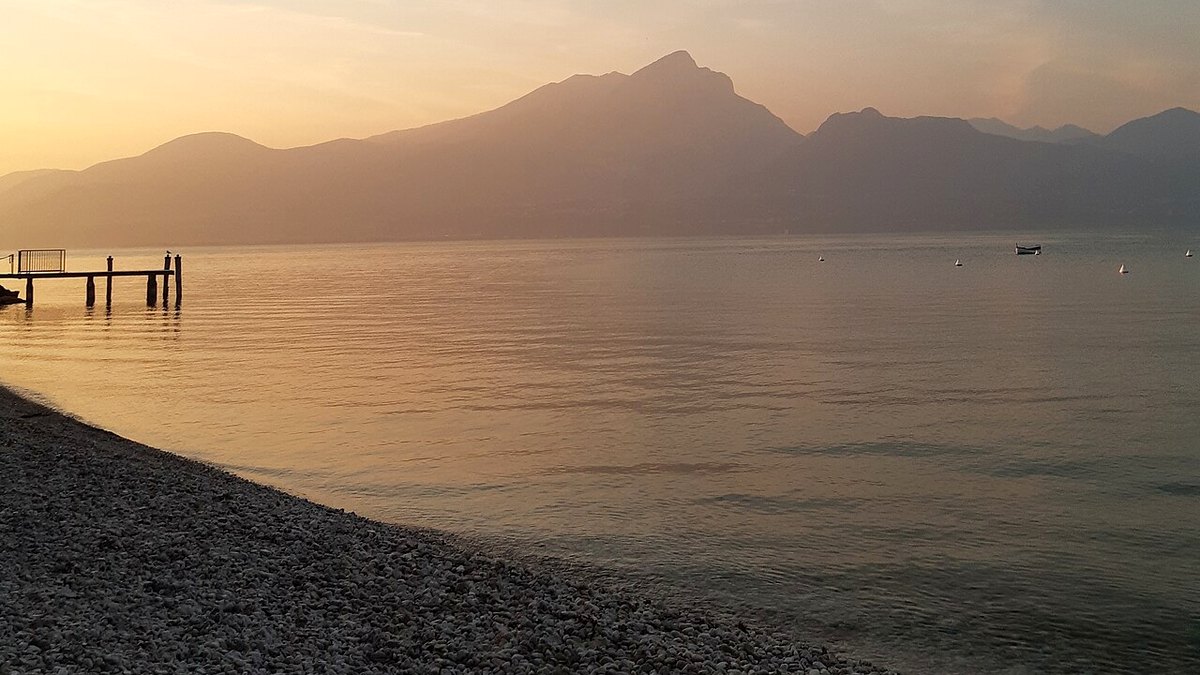 Kieselstrand in Torri del Benaco im Abendlicht, links ein Bootssteg, dahinter die Berge des Westufers